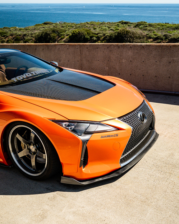 Front detail of Cassandra Balthasar’s orange widebody Lexus LC500 with carbon hood and ocean view in the background.