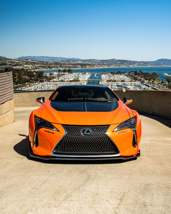 Front view of Cassandra Balthasar’s orange widebody Lexus LC500 overlooking a marina and coastal hillside.