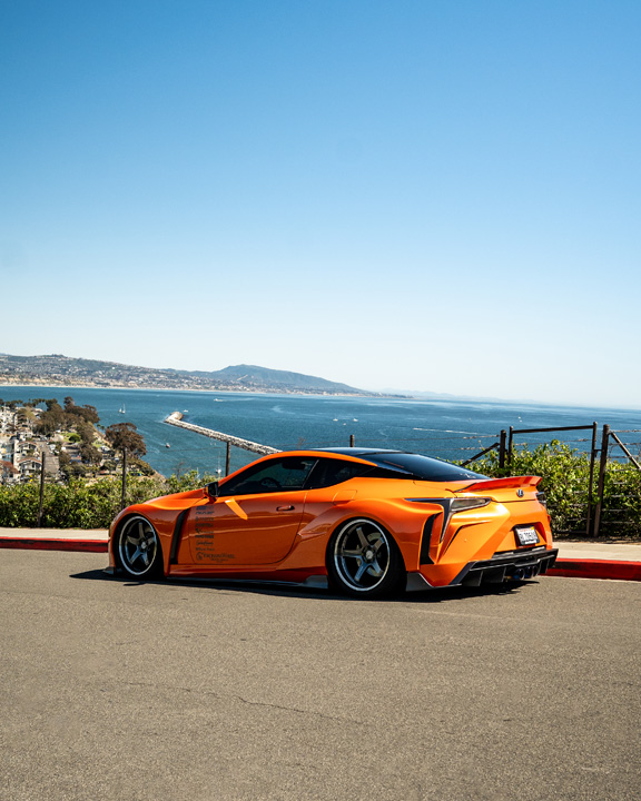 Cassandra Balthasar’s orange widebody Lexus LC500 parked along a coastal road overlooking the ocean.