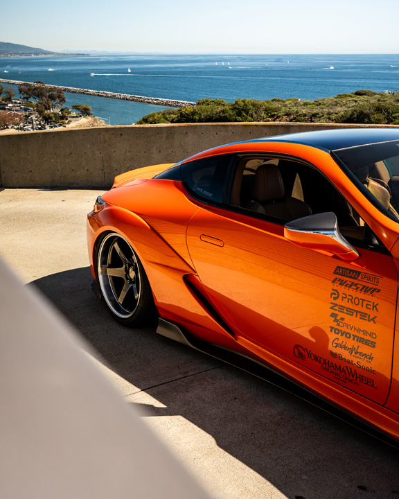 Side detail of Cassandra Balthasar’s orange widebody Lexus LC500 with ocean views and sponsor decals.