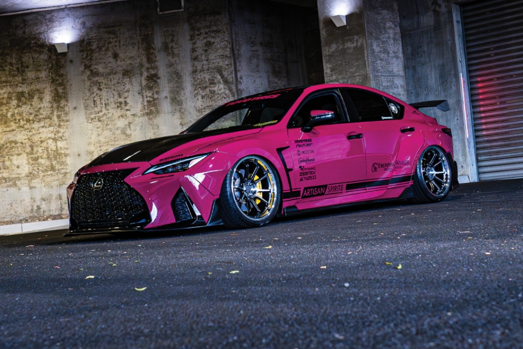 Giuseppe Balthasar’s pink 2023 Lexus IS 500 with an Artisan Spirits widebody kit parked under a concrete overhang at night.