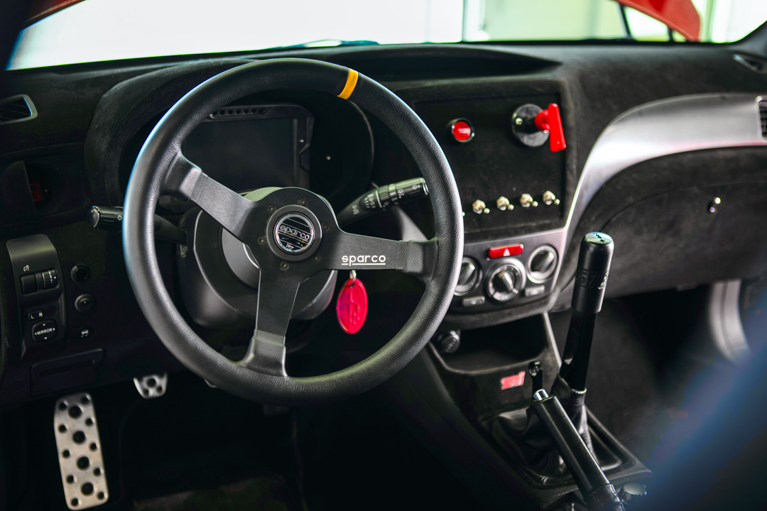 Interior view of Jon Erickson’s 2011 Subaru STi showing the Sparco steering wheel, flocked dash, custom switch panel, and shifter setup.