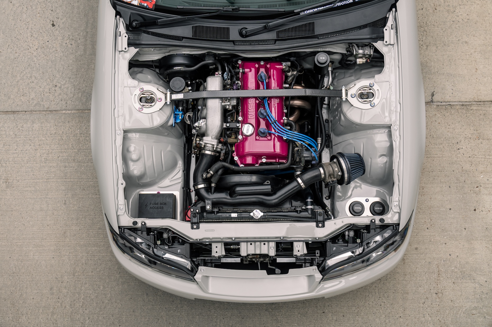 Overhead engine bay detail of Bill MacKenzie’s 2000 Nissan Silvia S15 showing the naturally aspirated SR setup, pink Nissan valve cover, strut bar, custom intake, and clean shaved bay.