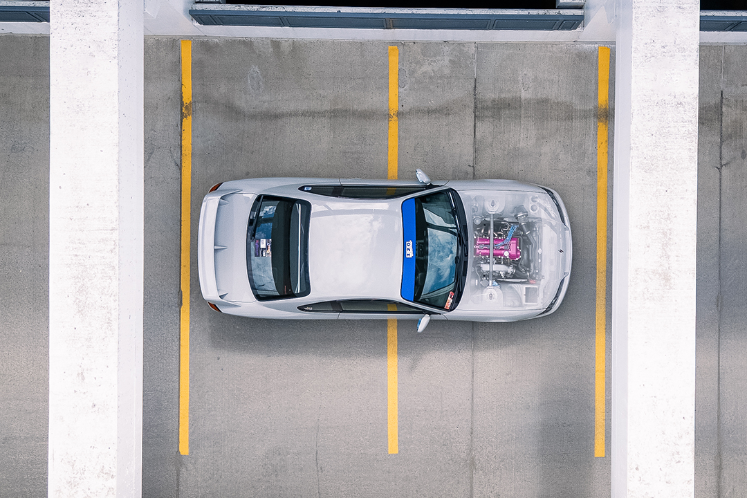 Overhead view of Bill MacKenzie’s 2000 Nissan Silvia S15 parked in a concrete garage, showing the clean body lines, blue windshield banner, and exposed engine bay through the front opening.