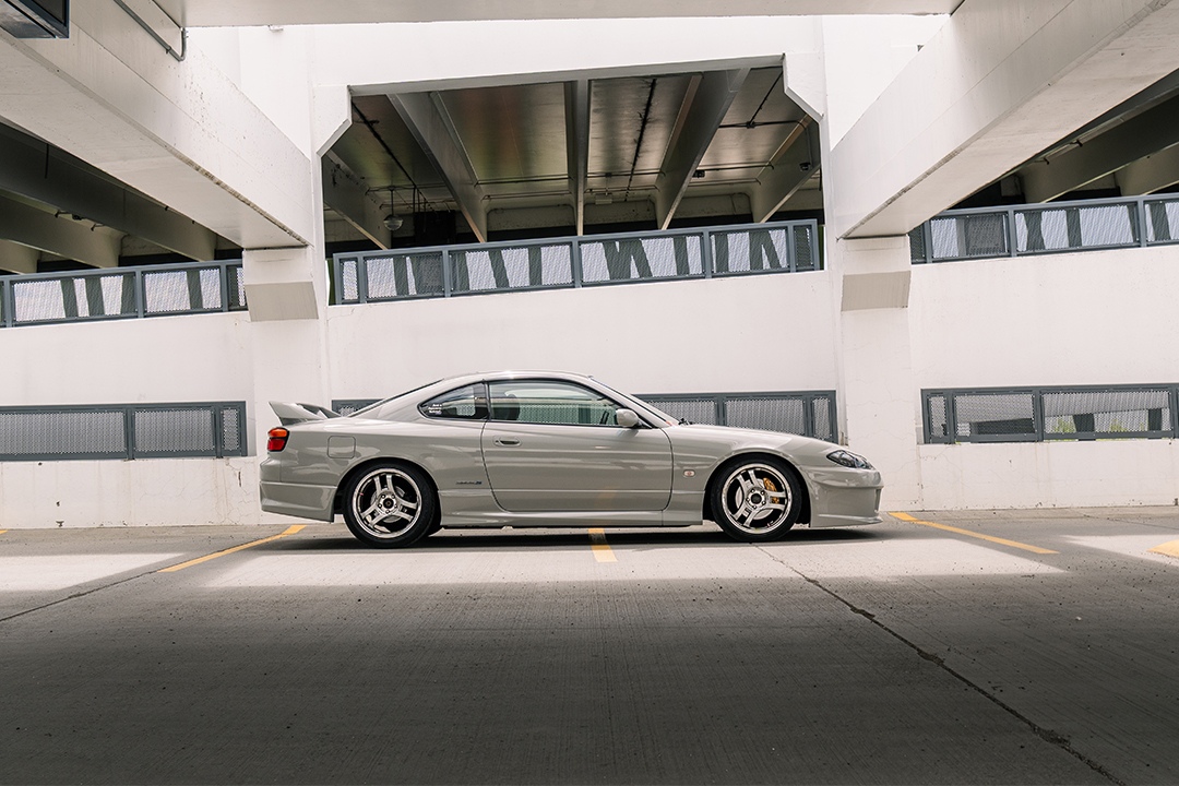 Side profile of Bill MacKenzie’s Porsche Chalk 2000 Nissan Silvia S15 parked in a concrete parking garage, showing the Nismo aero, lowered stance, and Super Advan V2 wheels.