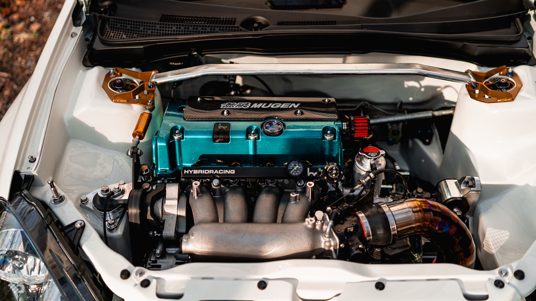 Engine bay detail of Moises Wynns’ 2003 Acura RSX showing the Skunk2-built K-series setup, Mugen valve cover, Hybrid Racing components, strut bar, and shaved bay.