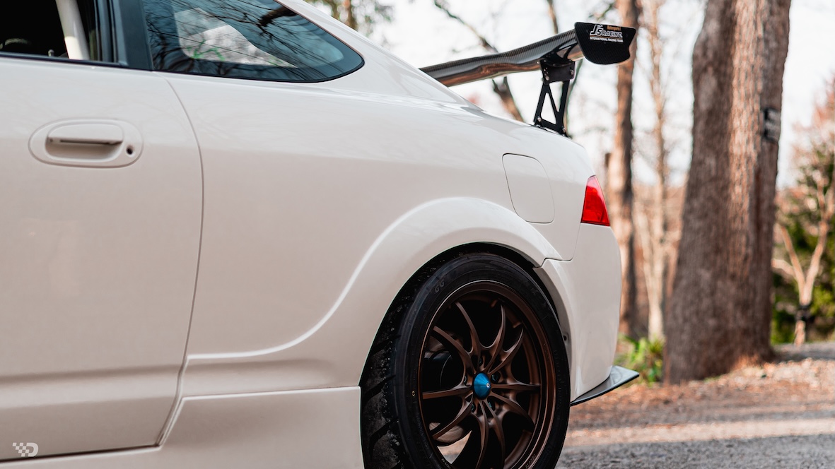 Rear quarter detail of Moises Wynns’ 2003 Acura RSX showing the molded rear fender flare, Mugen MF10 wheel, J’s Racing rear wing, and Championship White paint.