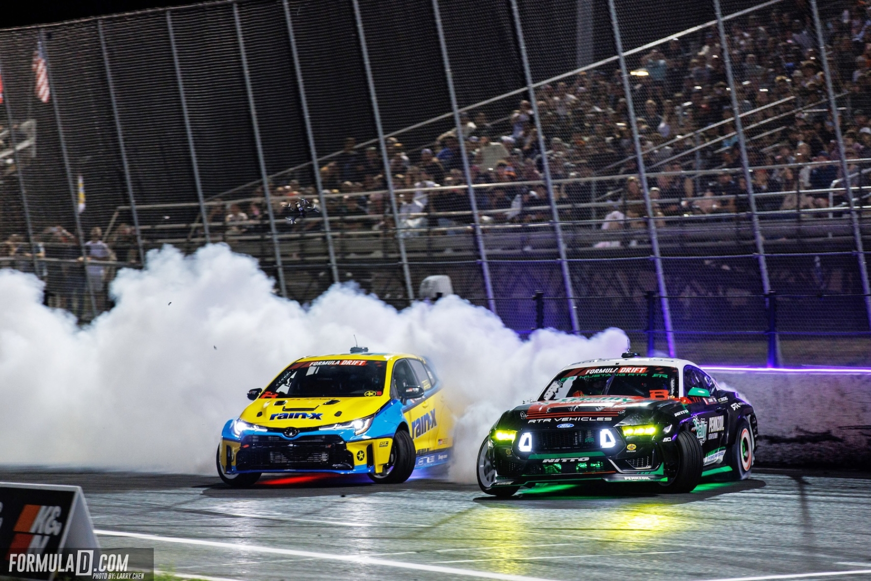 Two Formula DRIFT cars drifting at night in Long Beach with tire smoke, track lighting, and fans in the grandstands.