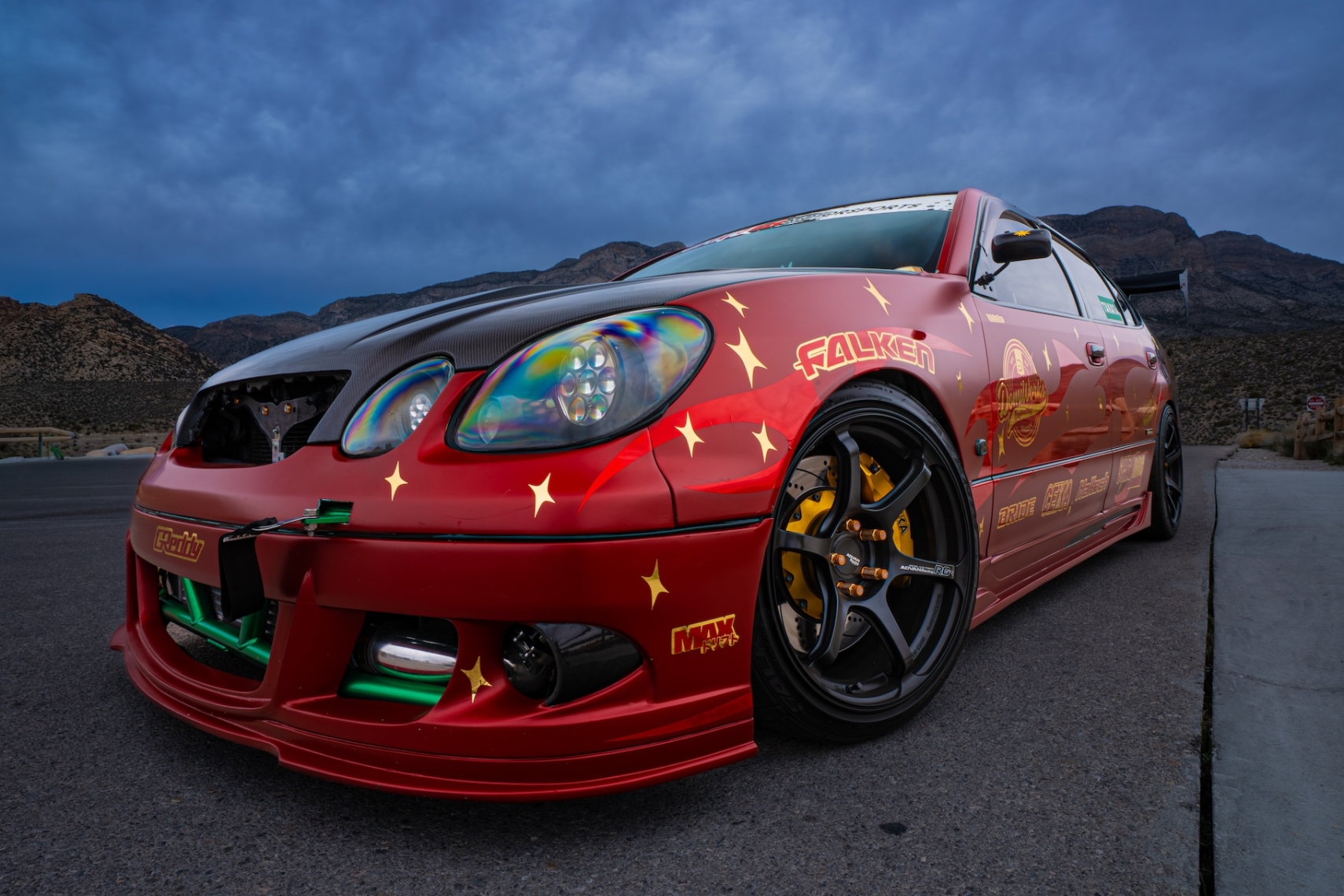 Front three-quarter view of Chris Burch’s satin red 1997 Toyota Aristo with RAW livery, carbon hood, Advan RG4 wheels, exposed front bash bar, and mountain backdrop.