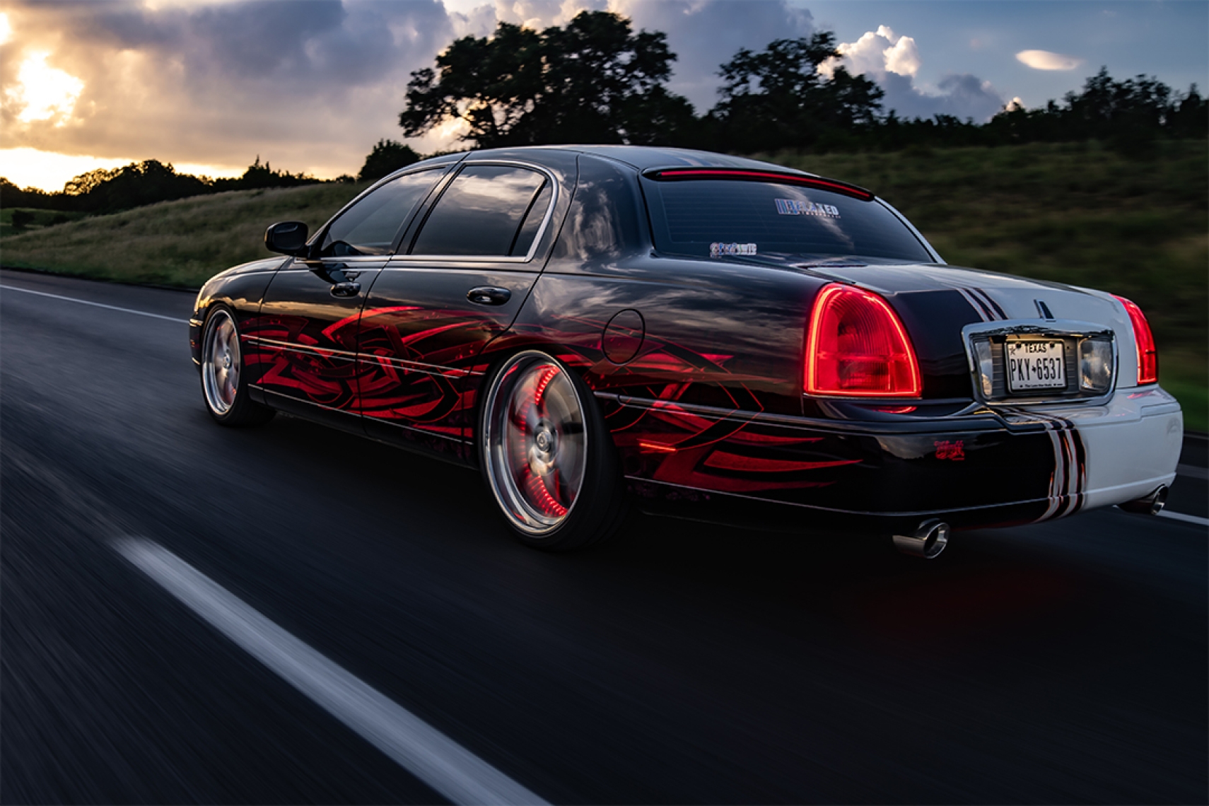 Rolling rear three-quarter view of Sam Benischek’s 2006 Lincoln Town Car showing the custom Two-Face graphic paint, low air suspension stance, KRNC forged wheels, chrome trim, and illuminated taillights.