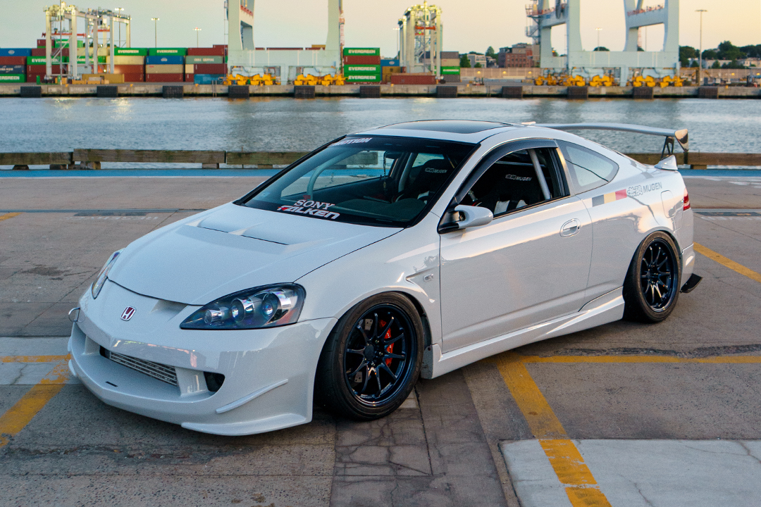 Front three-quarter view of Jonathan Ortiz’s 2005 Acura RSX Type-S showing the Porsche PTS Fashion Grey paint, Mugen front bumper and side skirts, J’s Racing hood, blue Volk Racing CE28N wheels, and shipping port backdrop.