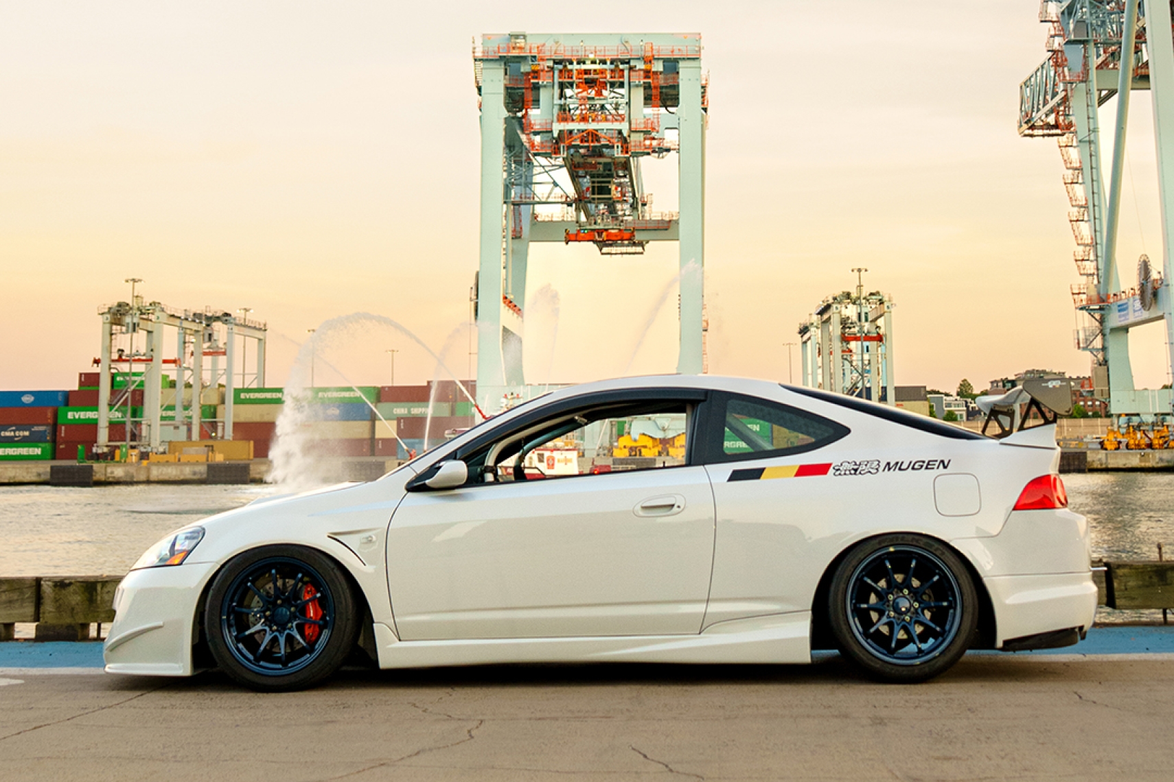 Side profile of Jonathan Ortiz’s 2005 Acura RSX Type-S showing the Porsche PTS Fashion Grey paint, Mugen aero, blue Volk Racing CE28N wheels, J’s Racing brake setup, and shipping port backdrop.