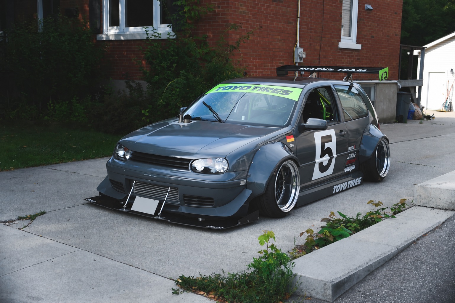 Current driveway photo of Jason Bos’ 2003 Volkswagen Golf GTI showing the handmade widebody, custom three-piece Enkei RPF1 wheels, Toyo Tires livery, front splitter, rear wing, and preserved show-car stance.