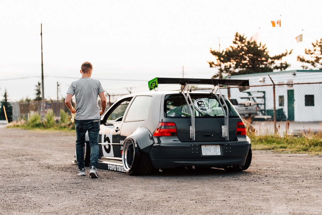 Rear three-quarter view of a widebody Volkswagen Golf show build with large rear wing and deep wheels, photographed with a person walking beside the car.