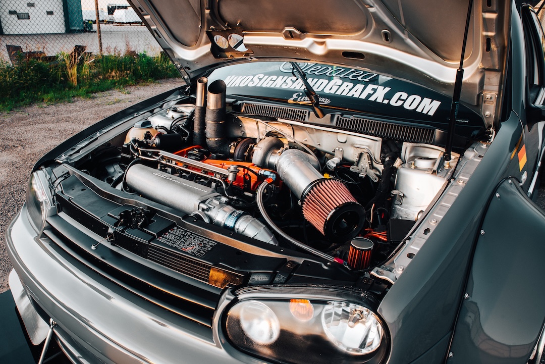 Turbocharged engine bay of a modified Volkswagen Golf with custom intake, exposed turbo setup, and widebody front-end details.