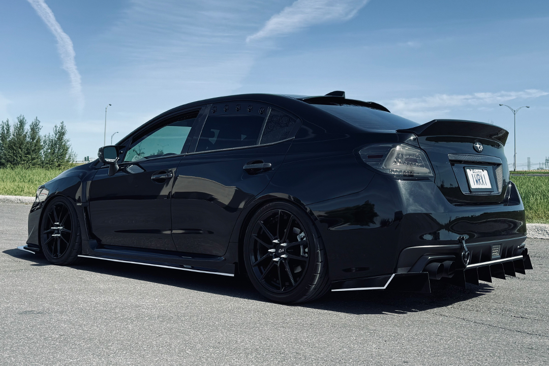 Rear three-quarter exterior view of Melissa Pelletier’s black 2017 Subaru WRX showing the black-and-white theme, gloss black wheels, ADE Creations aero, rear diffuser, window vents, duckbill spoiler, and lowered stance.