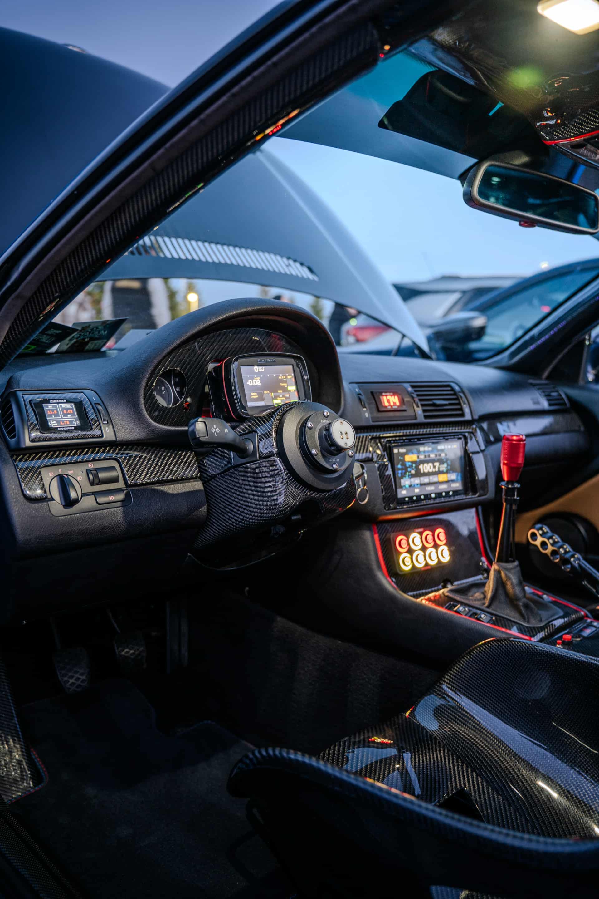 Interior of Jonathan St Pierre’s 2005 BMW 330Ci showing the FuelTech display and switch panel, carbon fiber trim, and Alpine head unit.